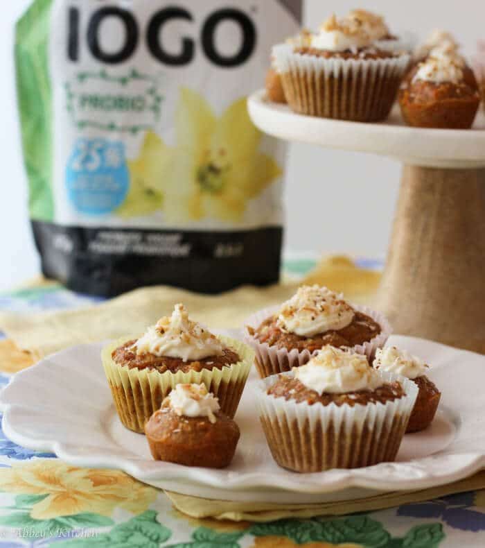 A plate with carrot cake cupcakes on a plate with a cake stand with more cupcakes on it behind it alongside a bag of yogurt.