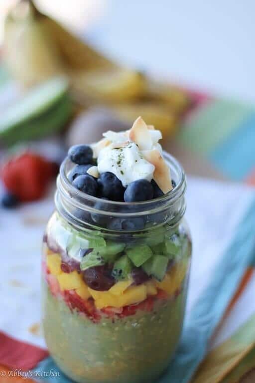 close up of vegan matcha overnight oatmeal in a clear jar garnished with fresh fruit, yogurt, and coconut flakes