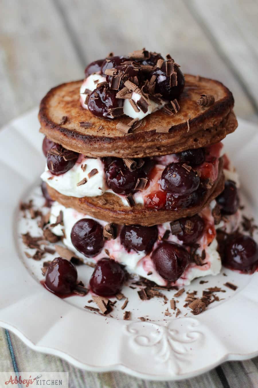 close up of a stack of chocolate protein pancakes served with cherries, whipped cream, and chocolate shavings on a white plate