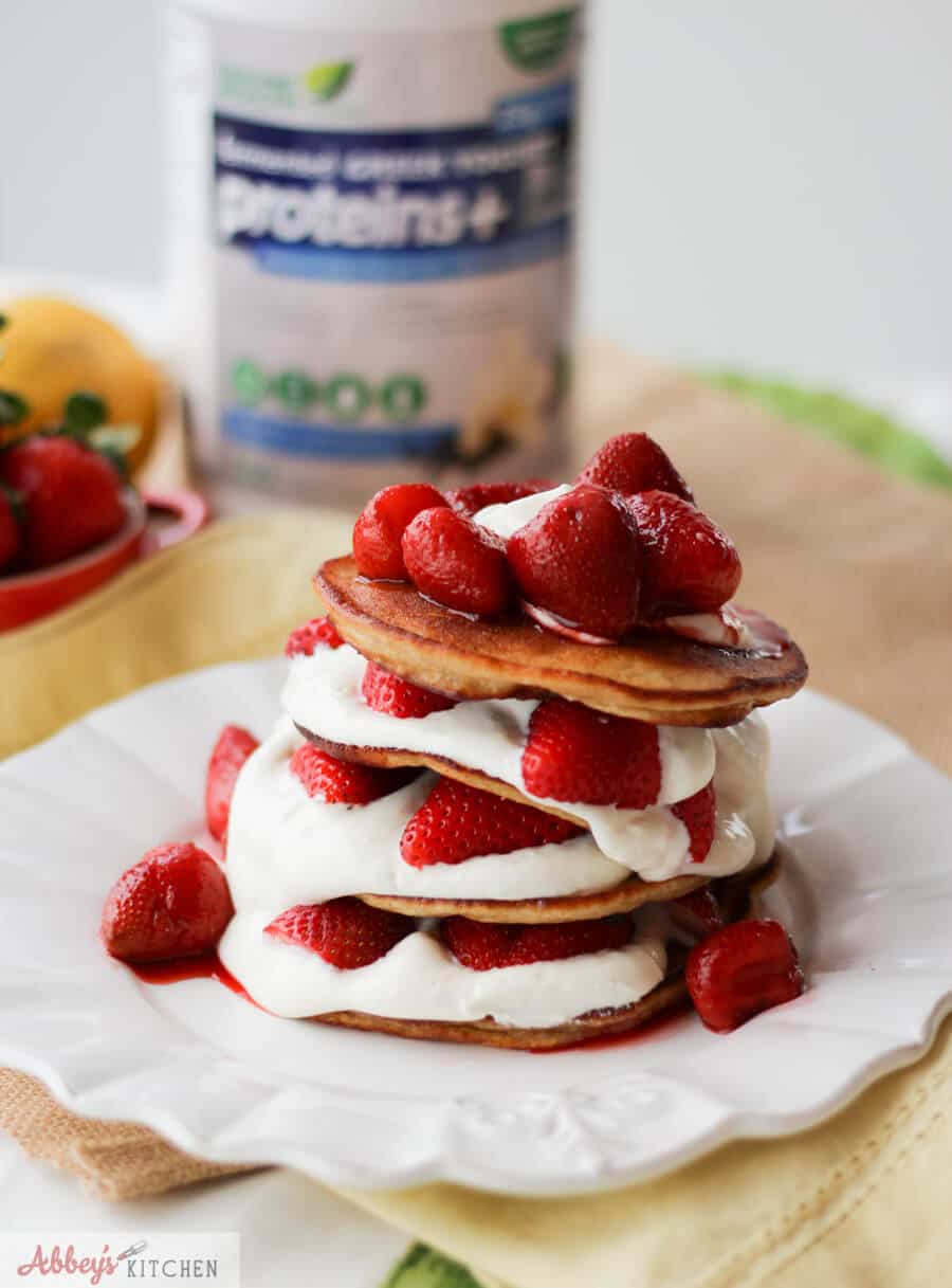 A photo of a stack of strawberry cheesecake protein pancakes with berries on top with a bottle of protein powder in the back.