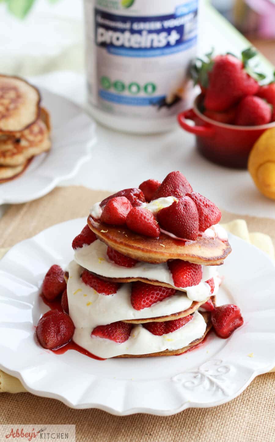 A stack of cheesecake pancakes with strawberries on top on a white plate with a bottle of protein powder in the back.