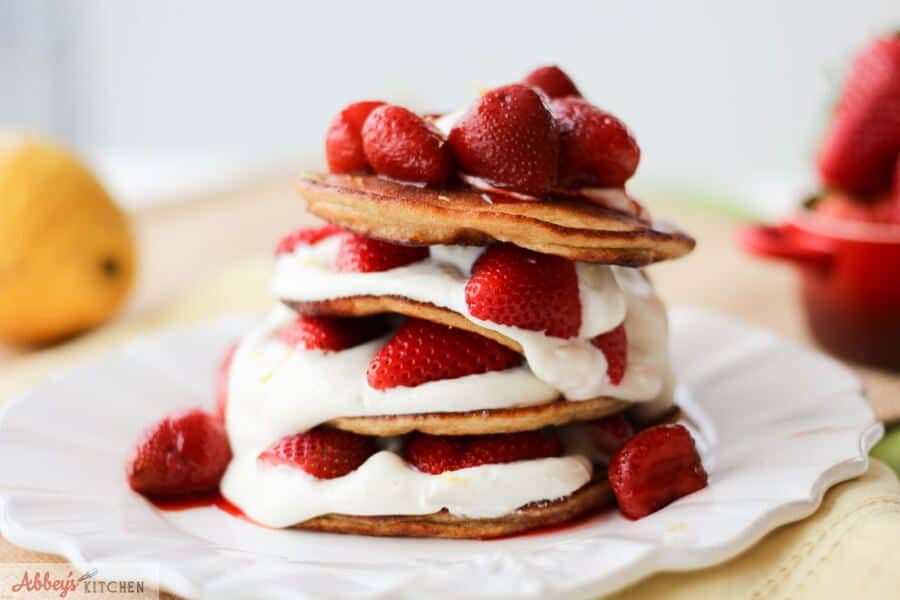 A stack of cheesecake pancakes with strawberries on top on a white plate.
