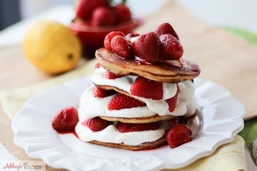 A stack of cheesecake pancakes with strawberries on top and around the pancakes on a white plate.