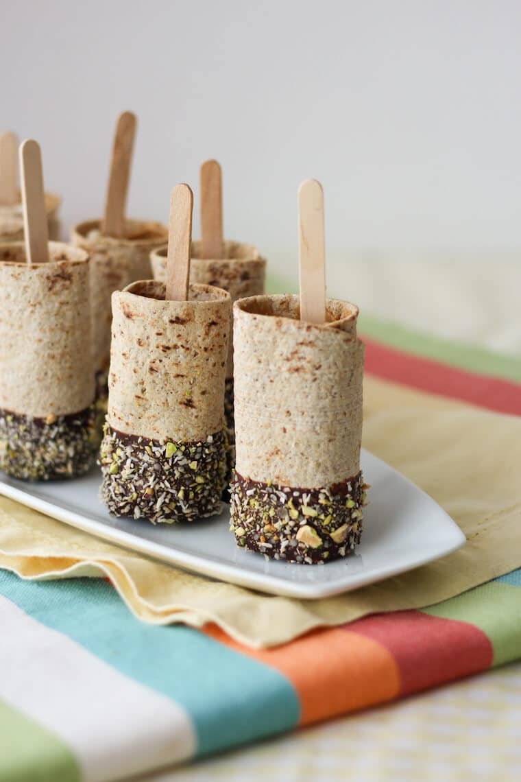 Photo of a plate of multiple vegan chocolate dipped banana roll ups on a stick.