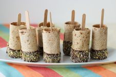 Photo of a plate of multiple vegan chocolate dipped banana roll ups on a stick.