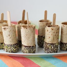 Photo of a plate of multiple vegan chocolate dipped banana roll ups on a stick.