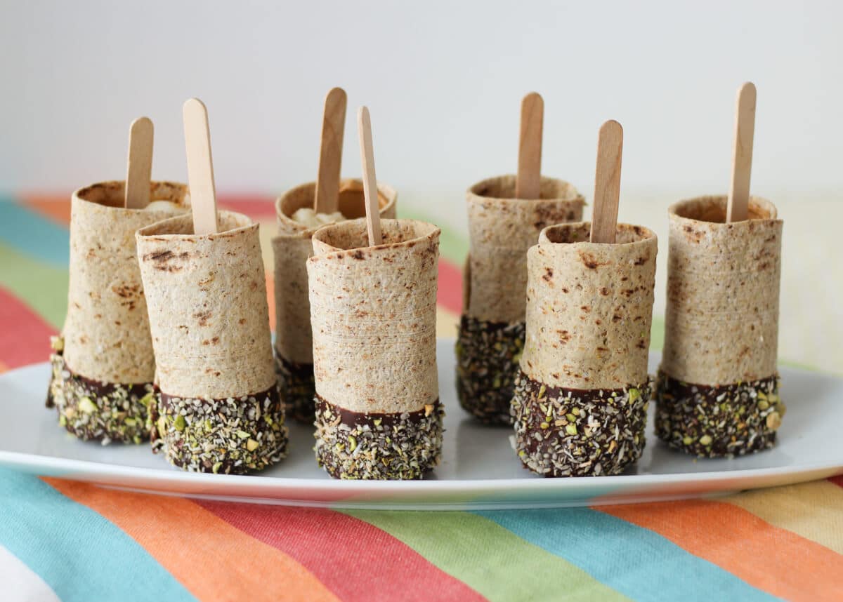 Photo of a plate of multiple vegan chocolate dipped banana roll ups on a stick.