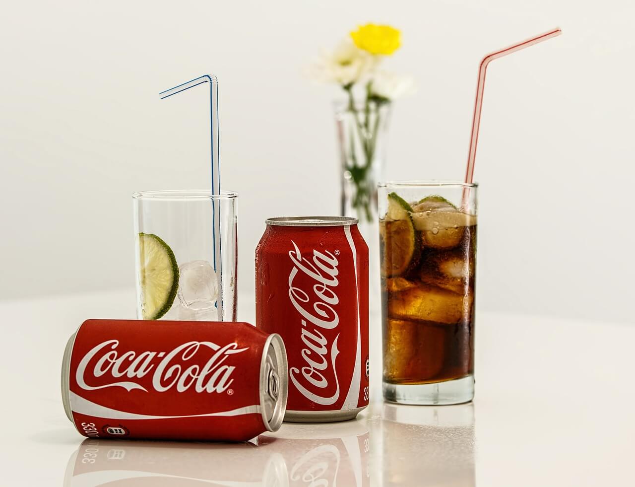 Two cans of coca cola beside two glasses with straws.