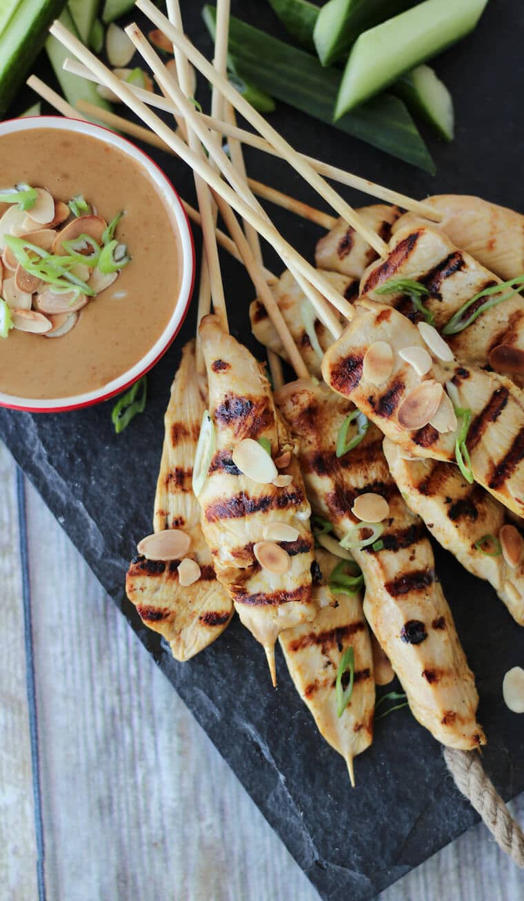 An overhead image of a serving platter of multiple chicken satay with thai almond sauce in the background.