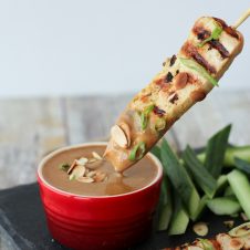 A skewer of chicken satay being dipped in an almond sauce.