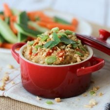 A red bowl of peanut hummus dip with sliced vegetables in the background.