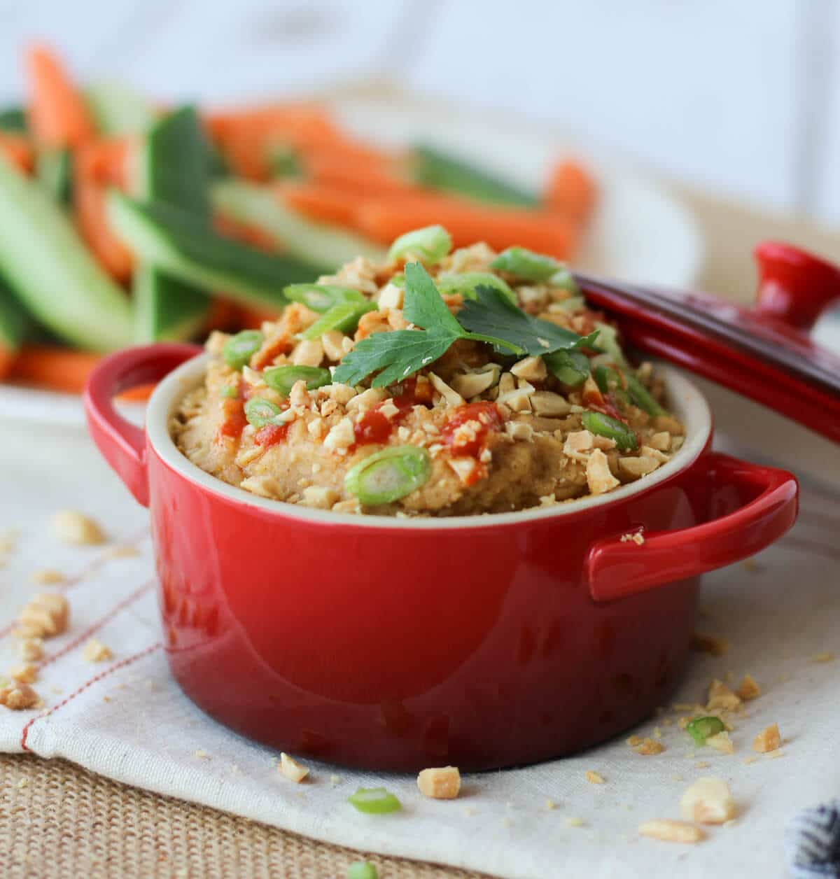 A red bowl of peanut hummus dip with sliced vegetables in the background.