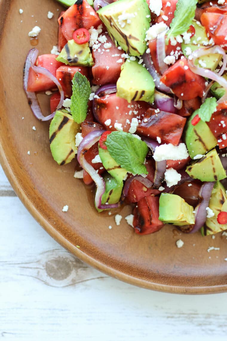 close up of grilled avocado and watermelon salad garnished with lime and feta on a wooden plate