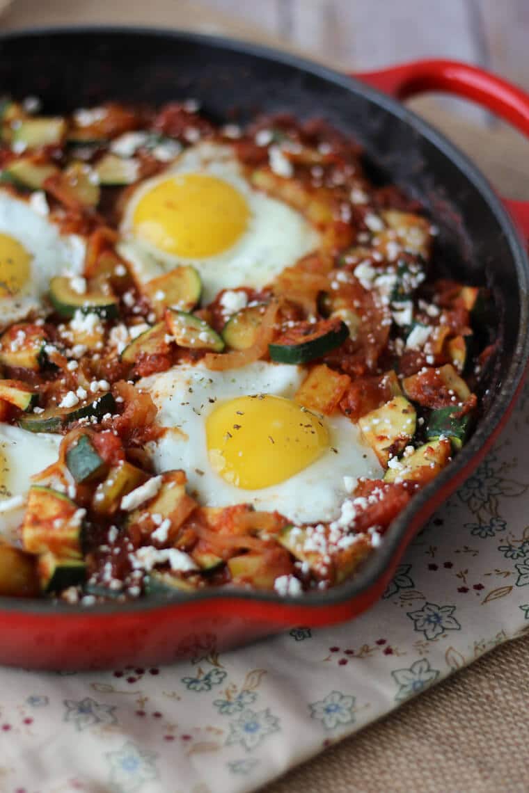 A close up photo of a red skillet containing a gluten free shaksuka with zucchini and feta.