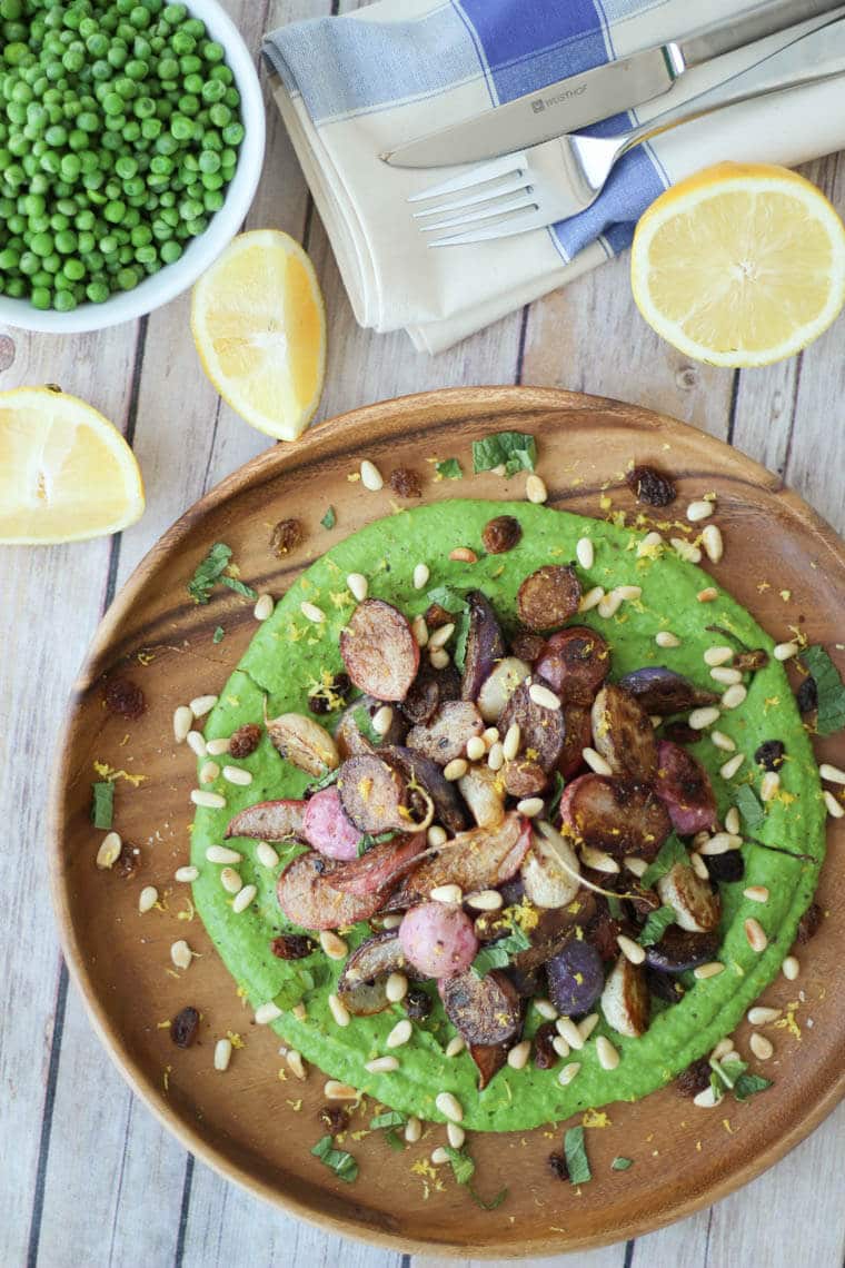 An overhead photo of a plate of za\'atar roasted radishes over top of a pea puree with sliced lemon and a bowl of peas around it.