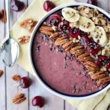 A white bowl containing a cherry smoothie with pecans, chia seeds, bananas, and cherries on top.