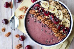 A white bowl containing a cherry smoothie with pecans, chia seeds, bananas, and cherries on top.