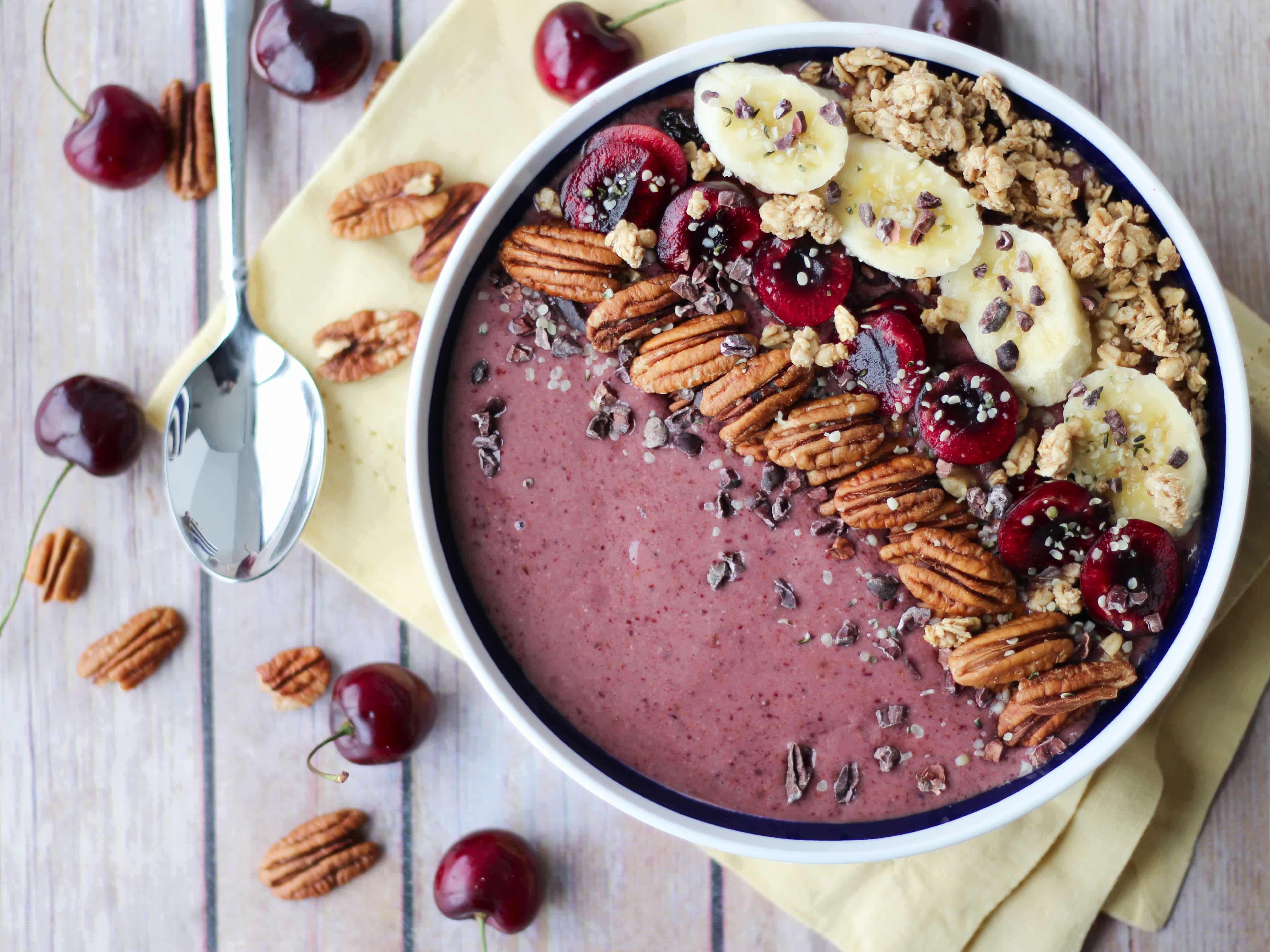 A white bowl containing a cherry smoothie with pecans, chia seeds, bananas, and cherries on top. 