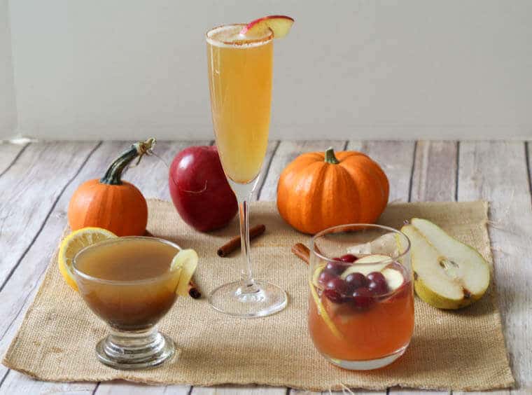 A table with three holiday cocktails with pumpkins, apples, and pear in the background.