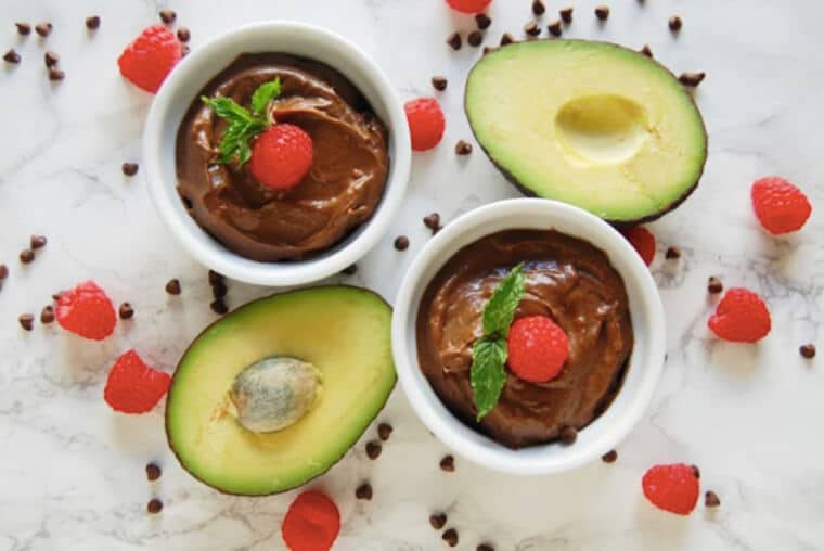 An overhead photo of two bowls of chocolate raspberry avocado pudding with avocados around it.