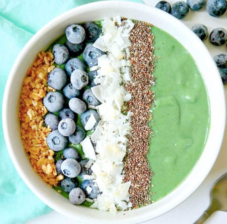 A smoothie bowl that is green and is topped with blueberries, coconut flakes, and chia seeds.