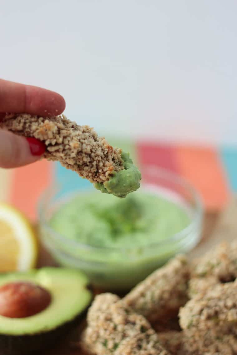 close up of an avocado fry dipped into green goddess dip with additional avocado fries and green goddess dip in a clear bowl in the background