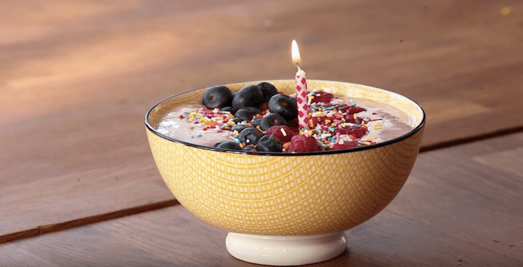 close up of a yellow bowl containing a smoothie bowl with a candle in it for celebrations from home