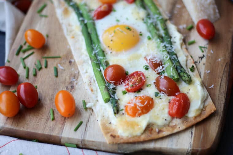 A breakfast pizza with egg, tomatoes, and asparagus on a wooden serving board. 