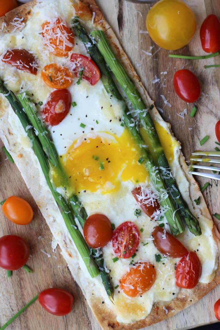 A breakfast pizza with egg, tomatoes, and asparagus with a fork beside it.