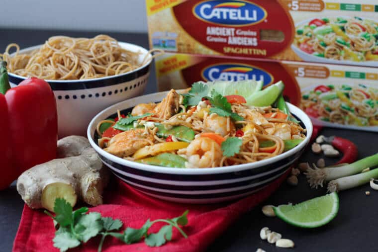 An angled image of a bowl of healthy lychee chicken curry noodles with garnish and limes on top with catelli pasta in a box in the background.
