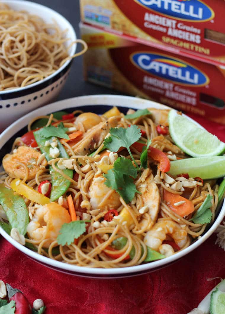 A bowl of healthy lychee chicken curry noodles with garnish and limes on top with boxed catelli pasta in the back.