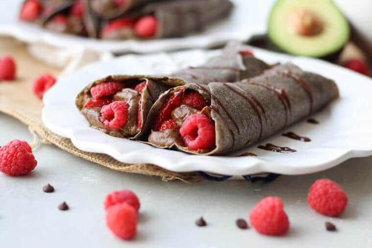 A white plate with two chocolate buckwheat crepes with raspberries inside and scattered around.