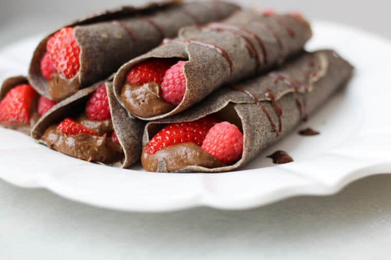A white plate of a stack of chocolate buckwheat crepes with raspberries.