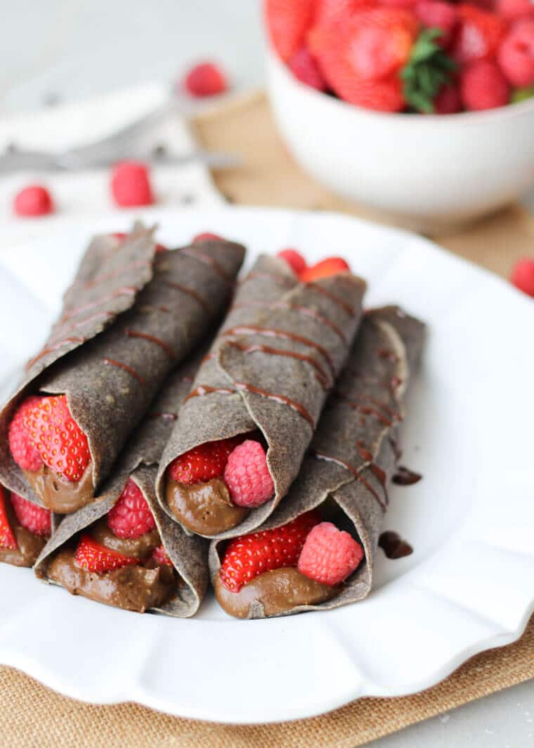 A white plate with a stack of multiple chocolate buckwheat crepes with raspberries.