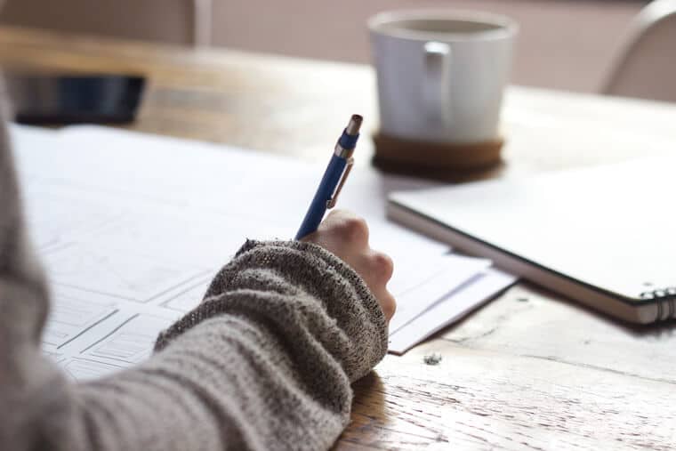 image of a person holding a pen doing work from home at a desk with a beverage in a mug