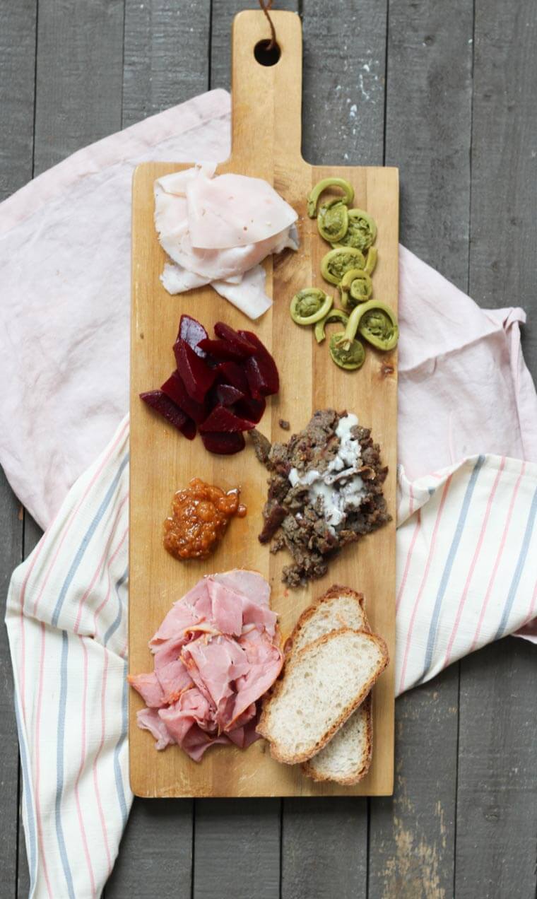 A wooden serving platter with charcuterie on it, such as cheese, sliced meats, bread slices, cut beets, and fiddleheads.