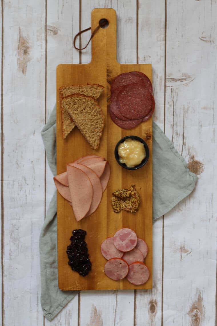 A wooden serving platter with charcuterie on it, such as cheese, sliced meats, bread slices, and nuts.