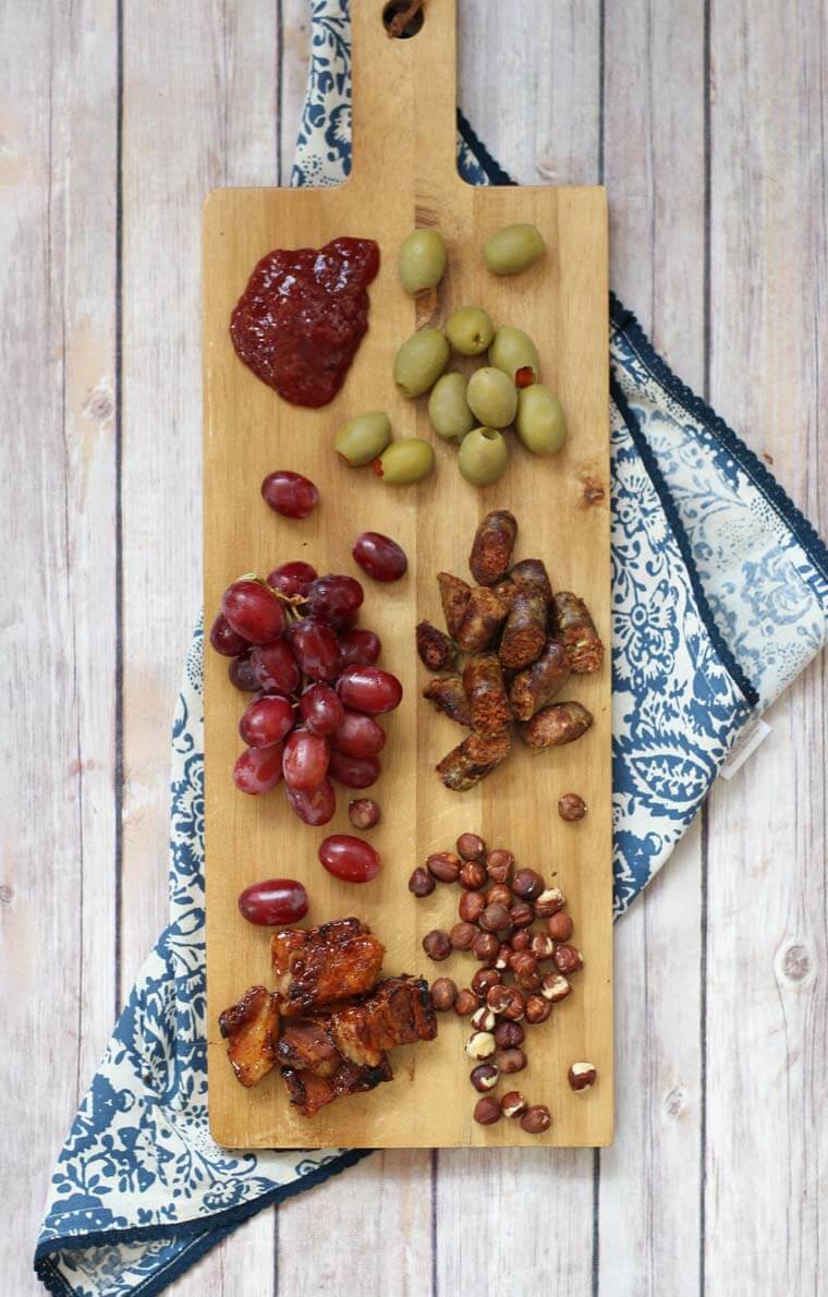 A wooden serving board with jam, olives, grapes, meat,  and nuts.