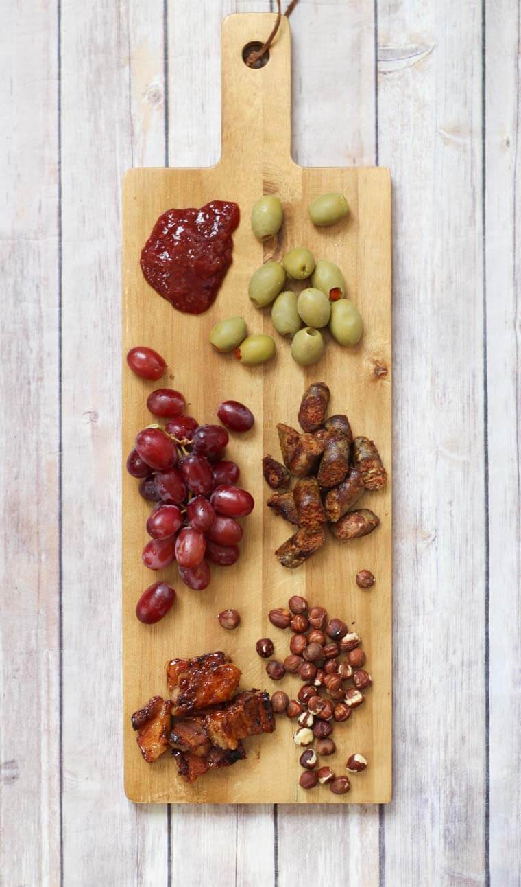 A wooden serving board with jam, olives, grapes, meat,  and nuts.