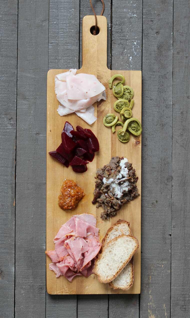 A wooden serving platter with charcuterie on it, such as cheese, sliced meats, bread slices, cut beets, and fiddleheads.