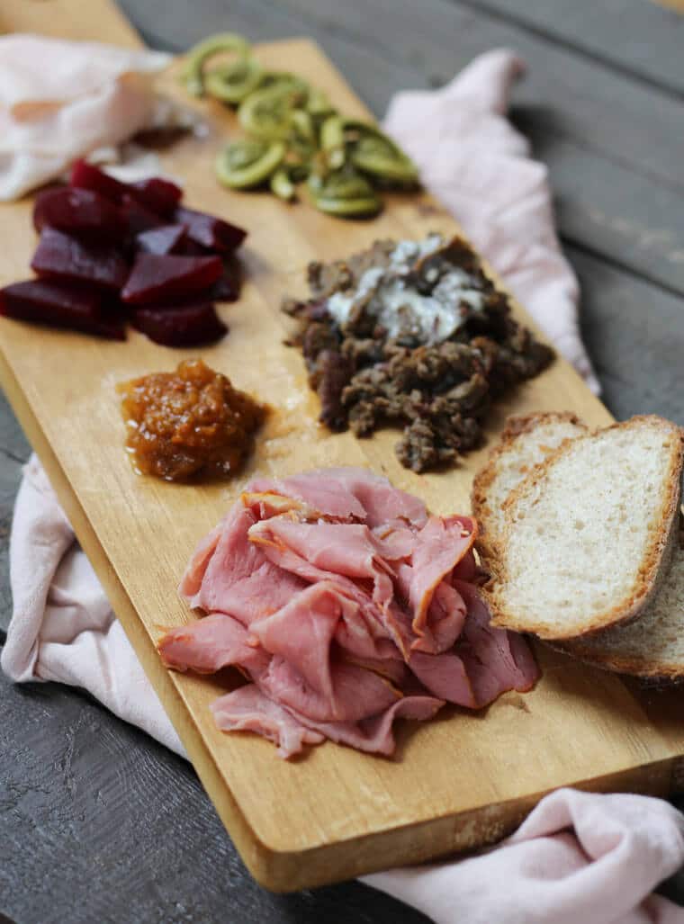 A wooden serving platter with charcuterie on it, such as cheese, sliced meats, bread slices, cut beets, and fiddleheads.