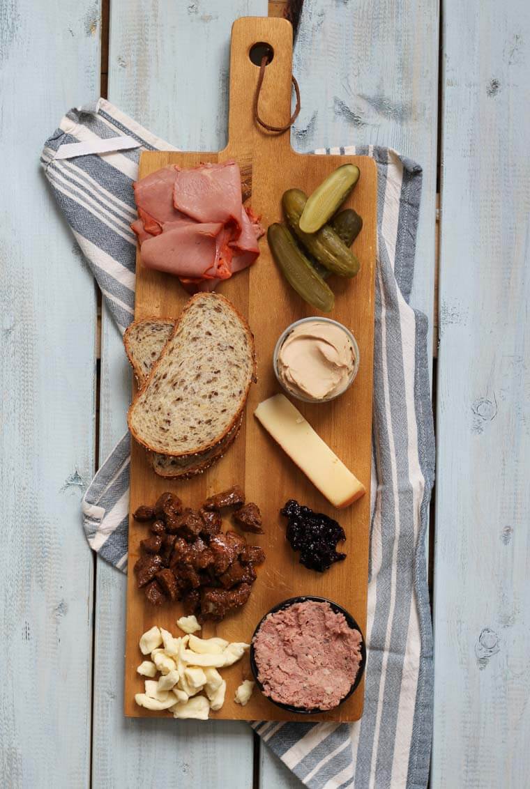 A wooden serving platter with charcuterie on it, such as cheese, meats, bread slices, pickles, and nuts.