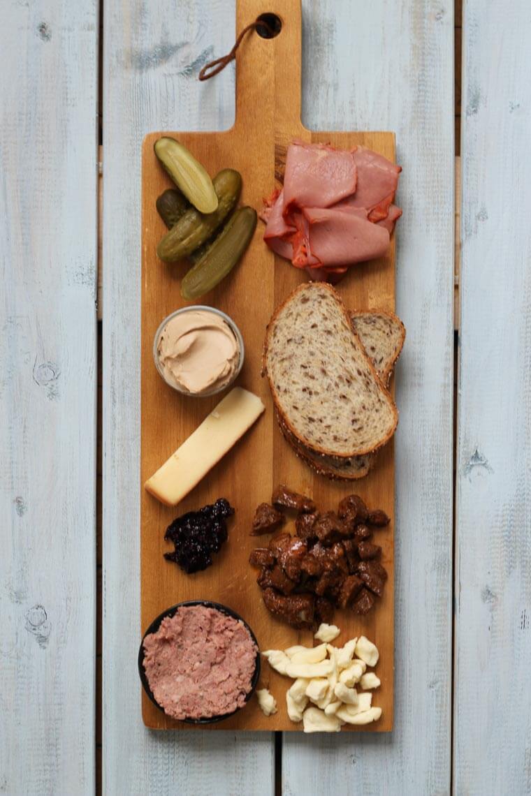 A wooden serving platter with charcuterie on it, such as cheese, meats, bread slices, pickles, and nuts.