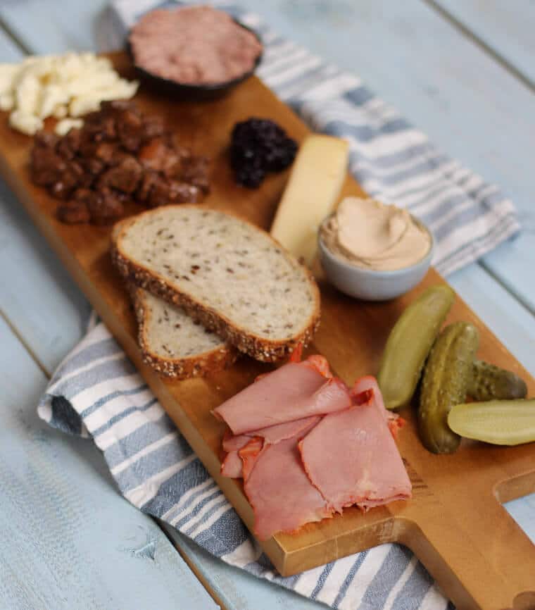 A wooden serving platter with charcuterie on it, such as cheese, meats, bread slices, pickles, and nuts.