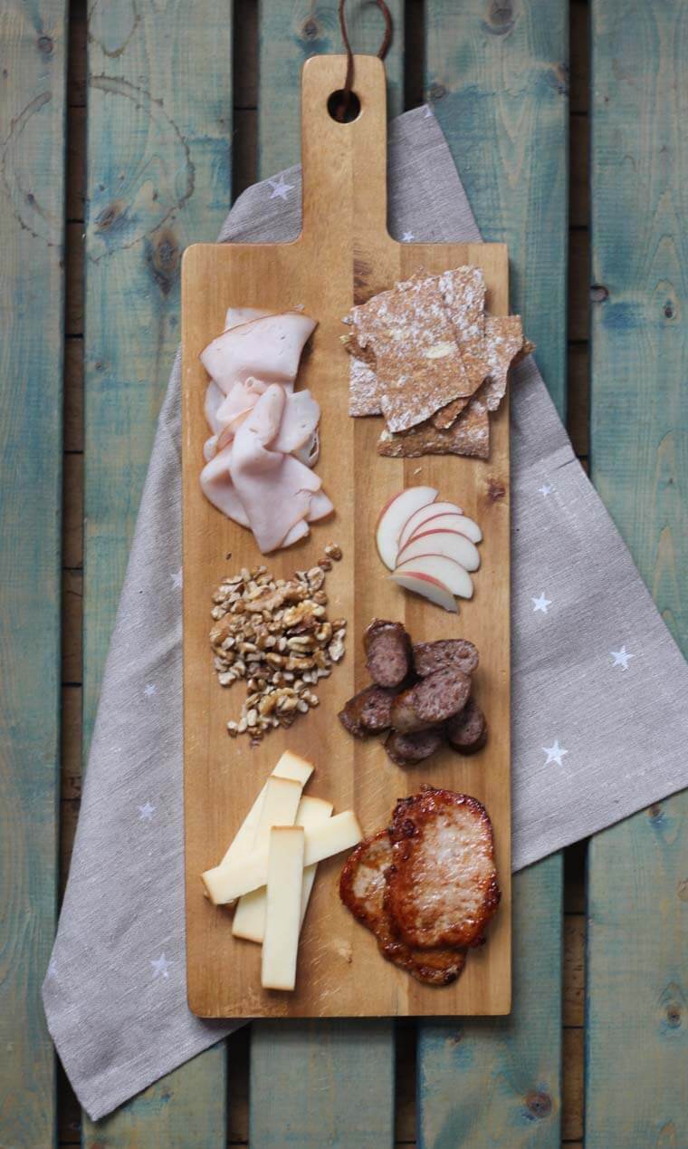 A wooden serving platter with charcuterie on it, such as cheese, meats, apple slices, and nuts.