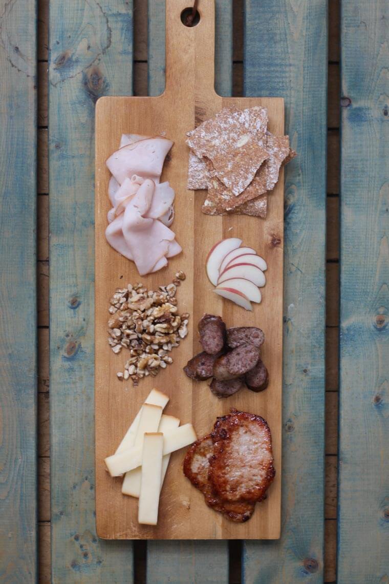 A wooden serving platter with charcuterie on it, such as cheese, meats, apple slices, and nuts.