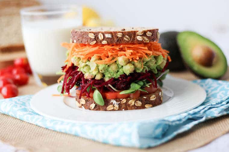 close up of vegan avocado and chickpea salad sandwich on brown bread served with fresh vegetables on a blue cloth