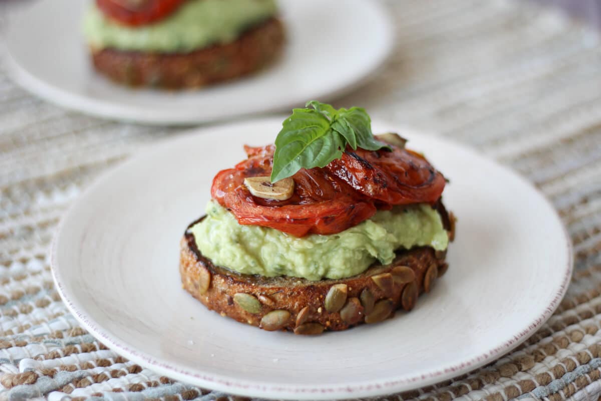 close up of a piece of toast topped with high protein avocado mixture, peppers, and fresh herbs on a white plate with additional piece of toast in the background