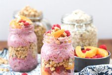 Two mason jars containing peach melba crumble overnight oats with a jar of oats and bowl of peaches in the background.