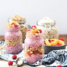 Two mason jars containing peach melba crumble overnight oats with a jar of oats and bowl of peaches in the background.